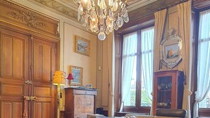 A luxurious living room with a chandelier, leather sofas, wooden flooring, and a large window with curtains. - Villa Primerose (Arcis-sur-Aube)