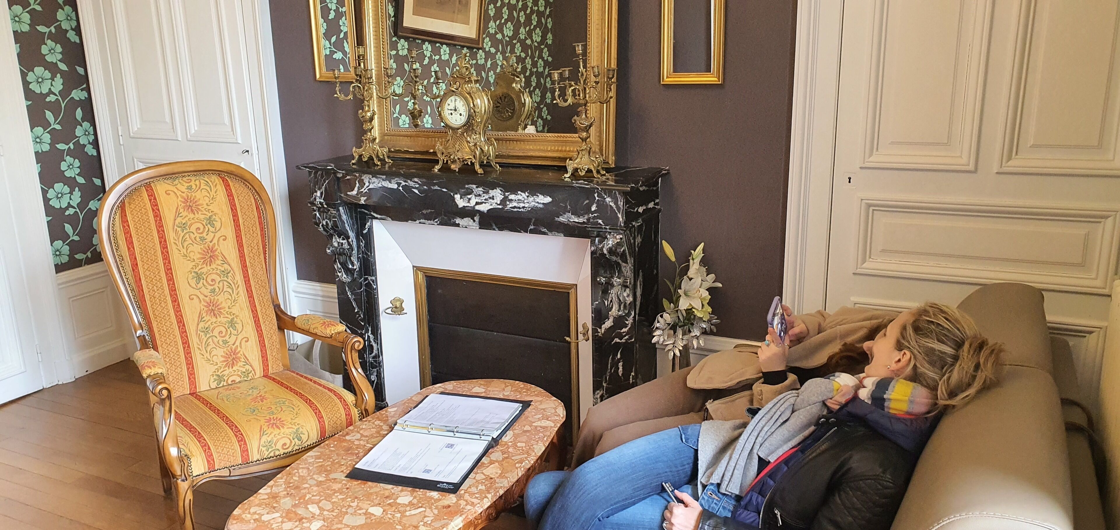A room with a fireplace, a mirror, a floral wallpaper, and a sofa.