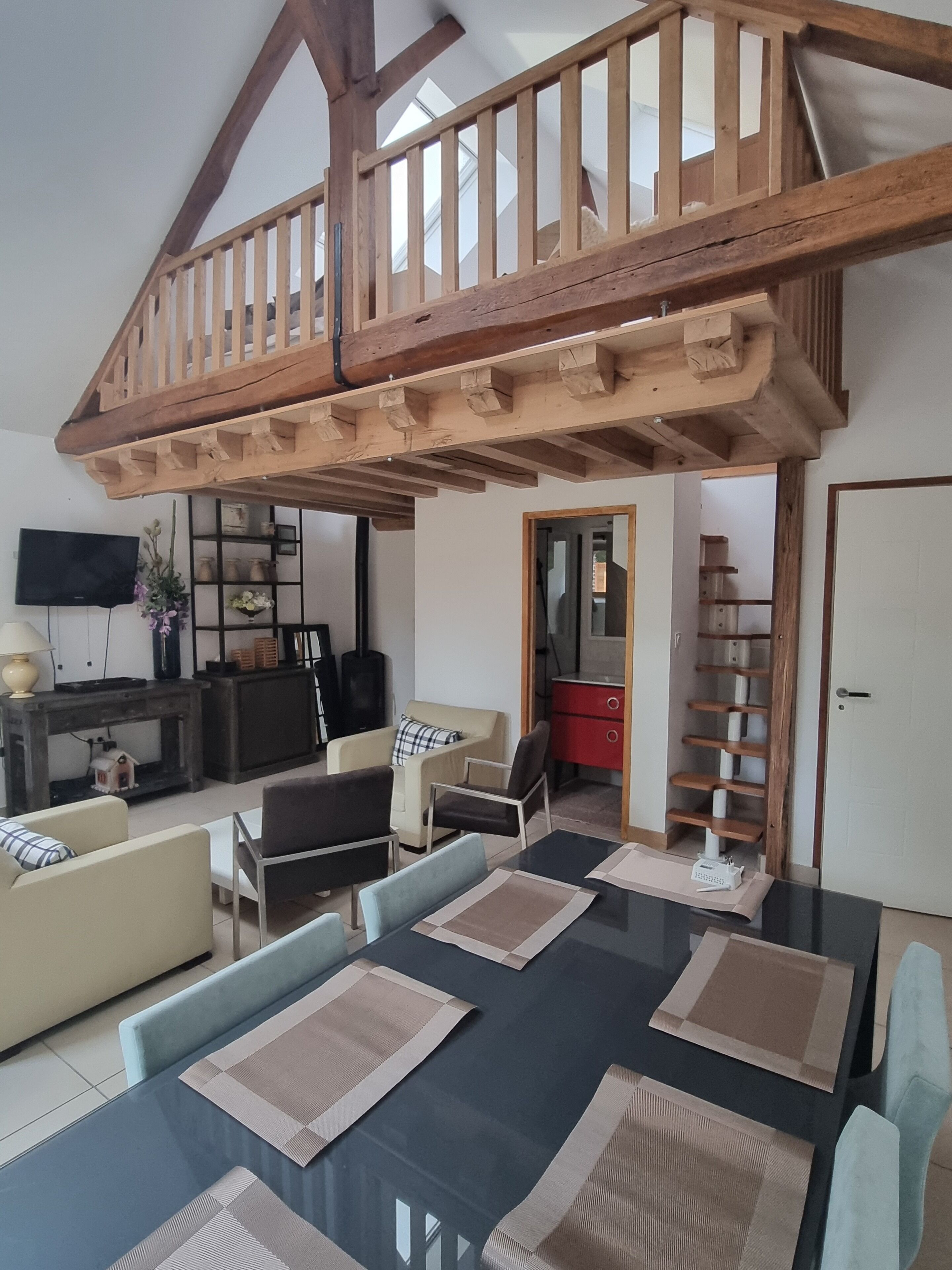 Ein geräumiges Wohnzimmer mit hoher Decke und freiliegenden Holzbalken, eine Essecke mit einem schwarzen Tisch und beigen Untersetzern sowie ein rotes Sideboard.