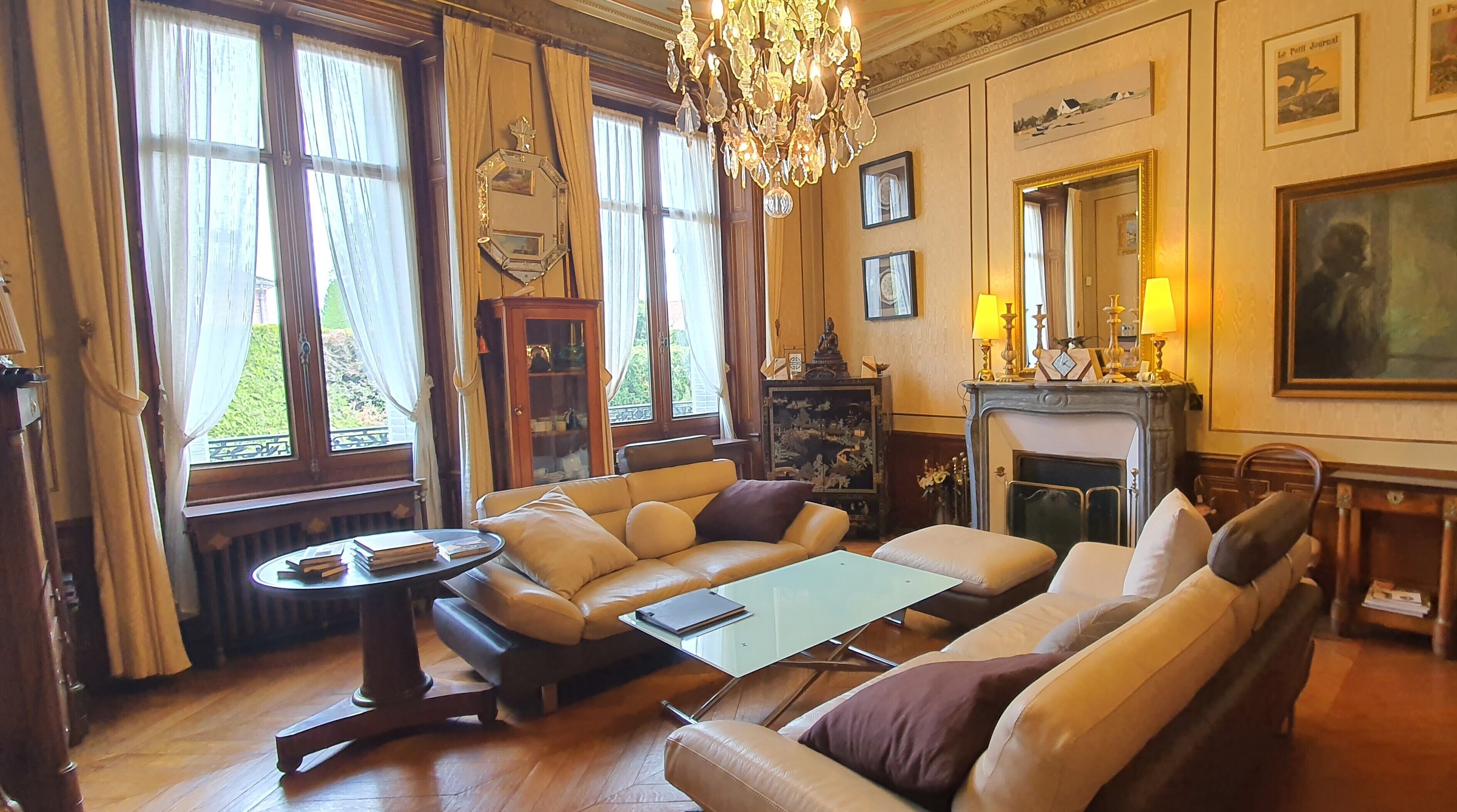 A spacious living room with a leather sofa, a glass coffee table, and a fireplace. There are large windows with curtains, a chandelier, and framed artwork on the walls.