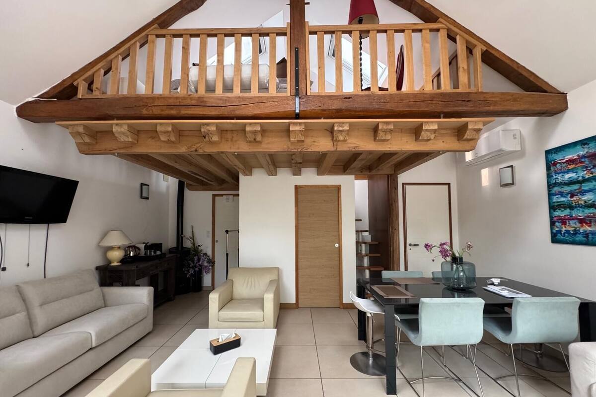Uma sala de estar ampla com pé direito alto, varanda de madeira, sofá, mesa de jantar com cadeiras e TV fixada na parede.