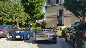 A parking area with several cars, including a blue convertible and a grey sedan, in front of a building with a stone facade and arched windows. - Villa Primerose (Arcis-sur-Aube)
