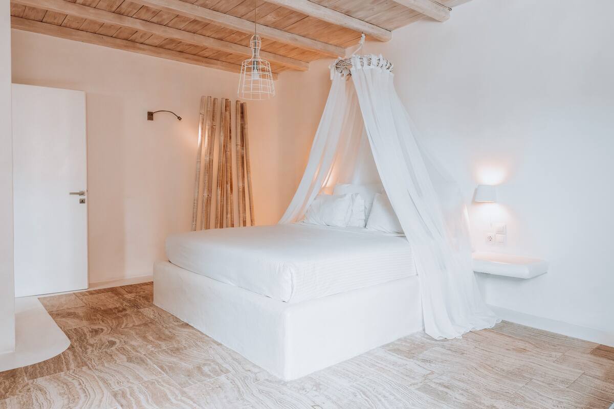 A bedroom with a bed, a bedside table, a nightstand, a wooden ceiling, and a wall-mounted light fixture.