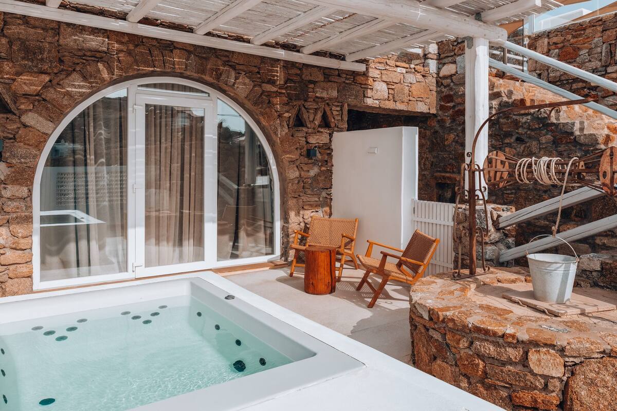 A stone-walled room with a glass door, a hot tub, wooden chairs, and a metal bucket.