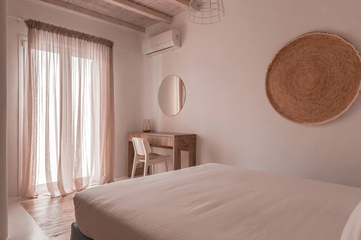 A bedroom with a bed, a desk, a chair, a round woven wall hanging, and a window with sheer curtains.
