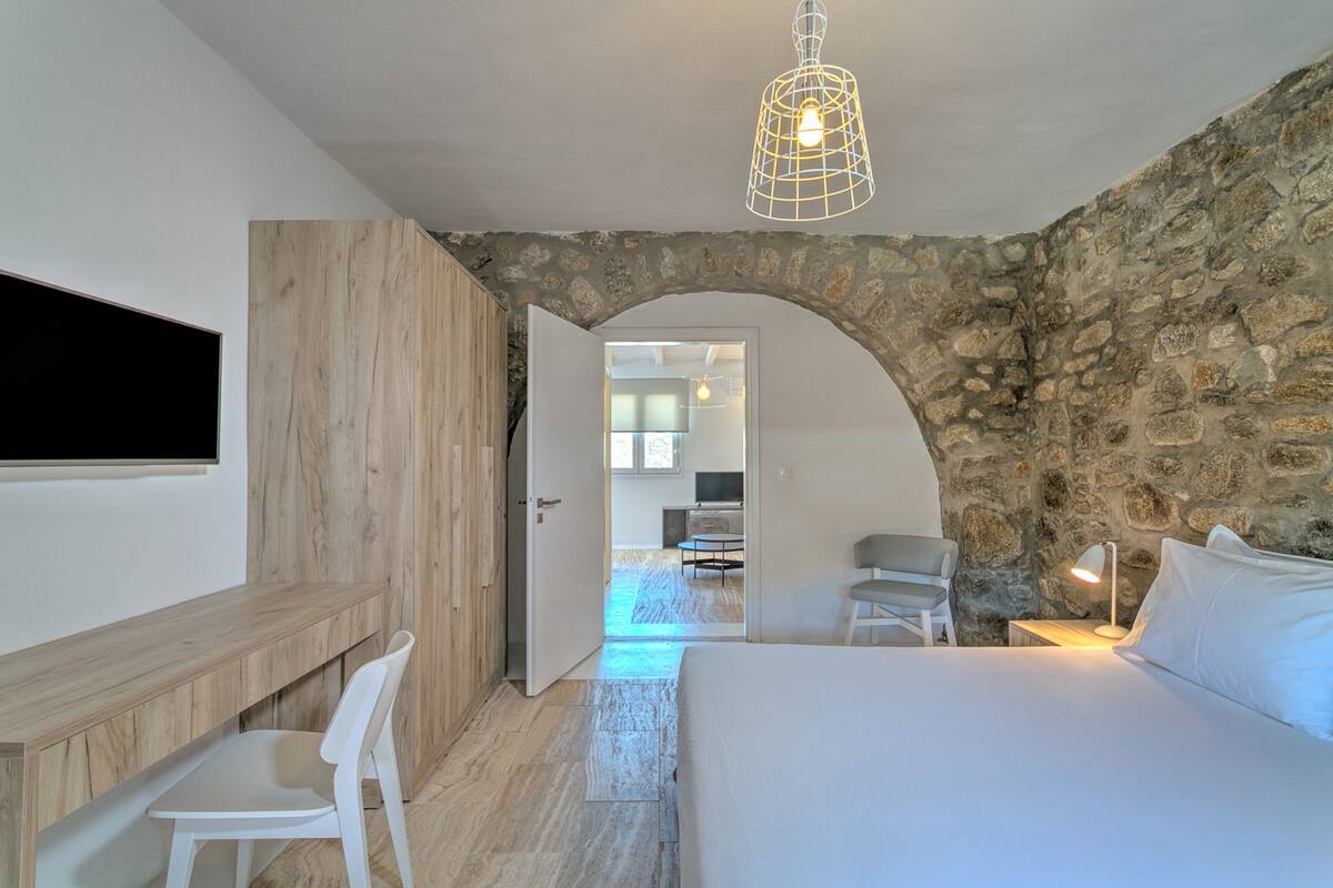 A bedroom with a stone wall, a wooden desk, a bed, and a TV mounted on the wall.