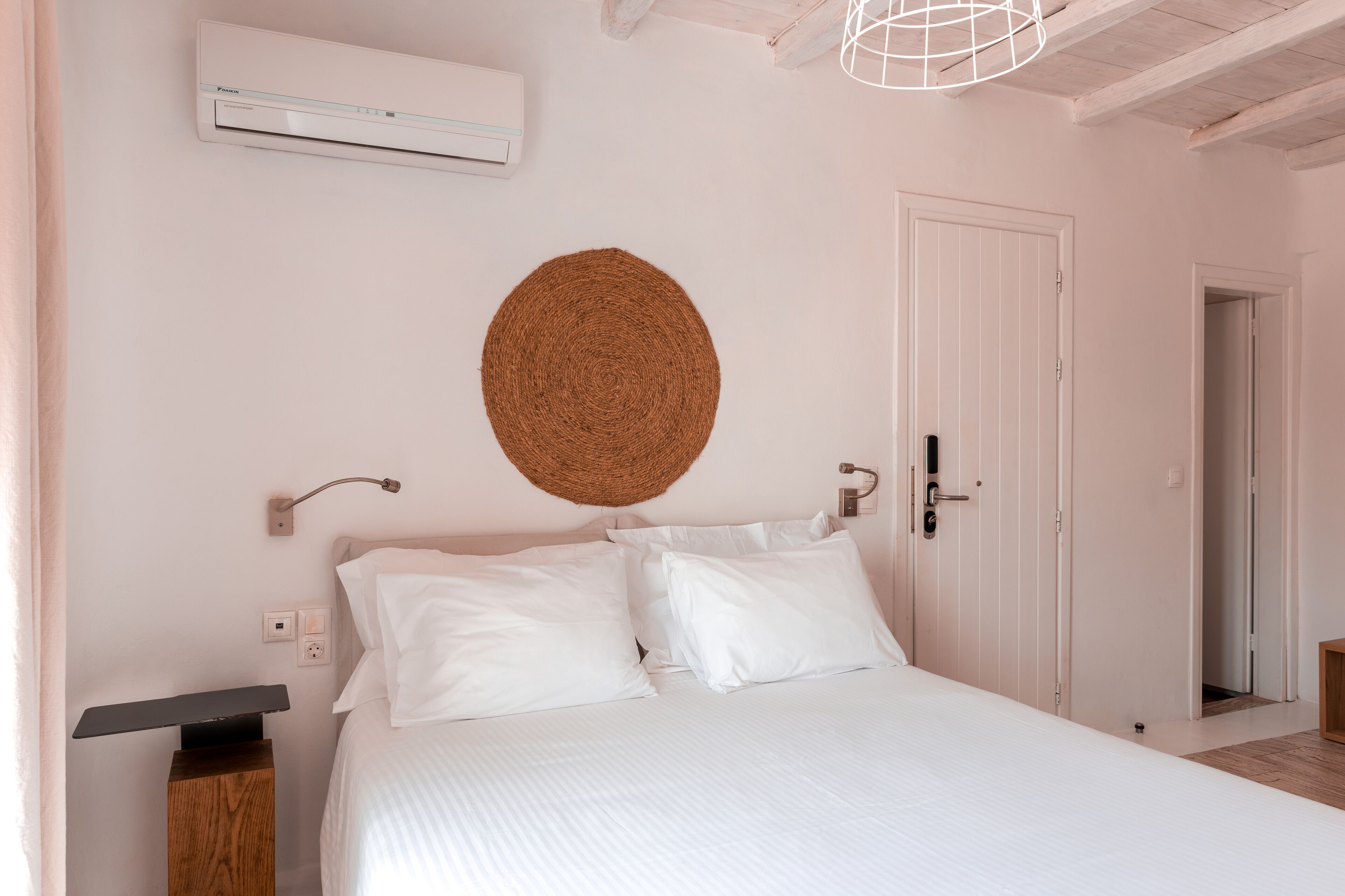 Une chambre à coucher avec un lit, des murs blancs, un ventilateur de plafond, une décoration murale en fibres tressées, une table de chevet et une porte.