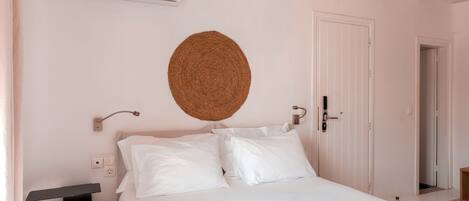 A bedroom with a bed, white walls, a ceiling fan, a woven wall hanging, a bedside table, and a door.