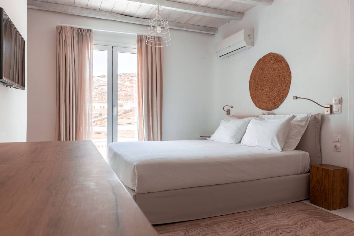 A bedroom with a bed, a wooden table, a woven wall hanging, and a view of the outdoors.