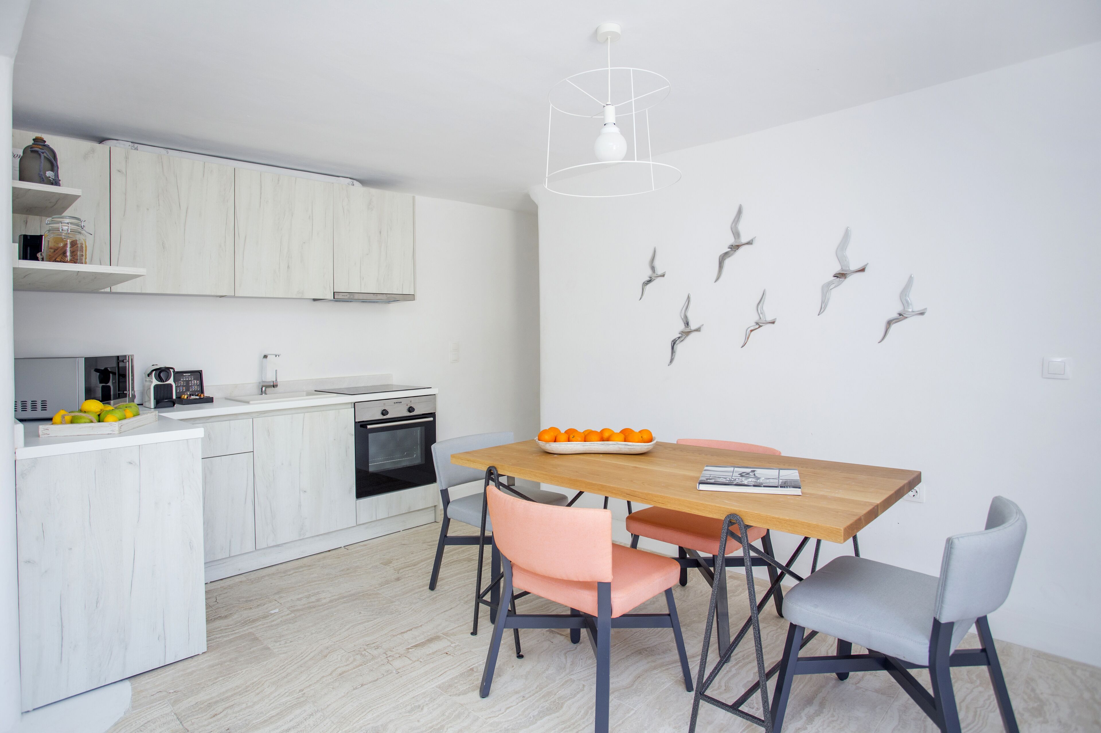 Una cocina moderna con armarios blancos, una mesa de comedor de madera y algunas sillas. Se observan adornos de pájaros en la pared y una lámpara colgante.