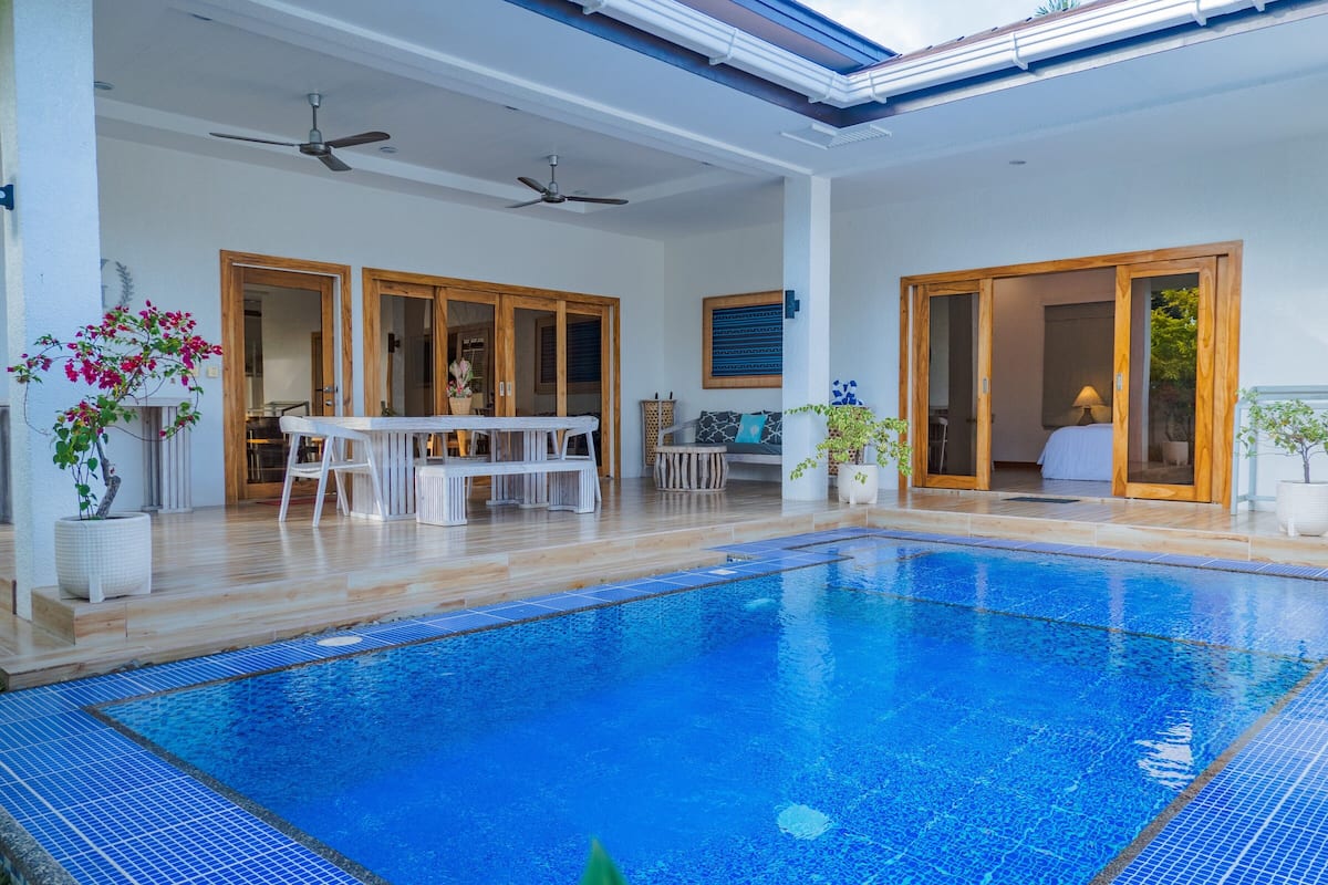 Une piscine intérieure spacieuse avec une terrasse en bois, un coin repas et une chambre visible à travers des portes vitrées.