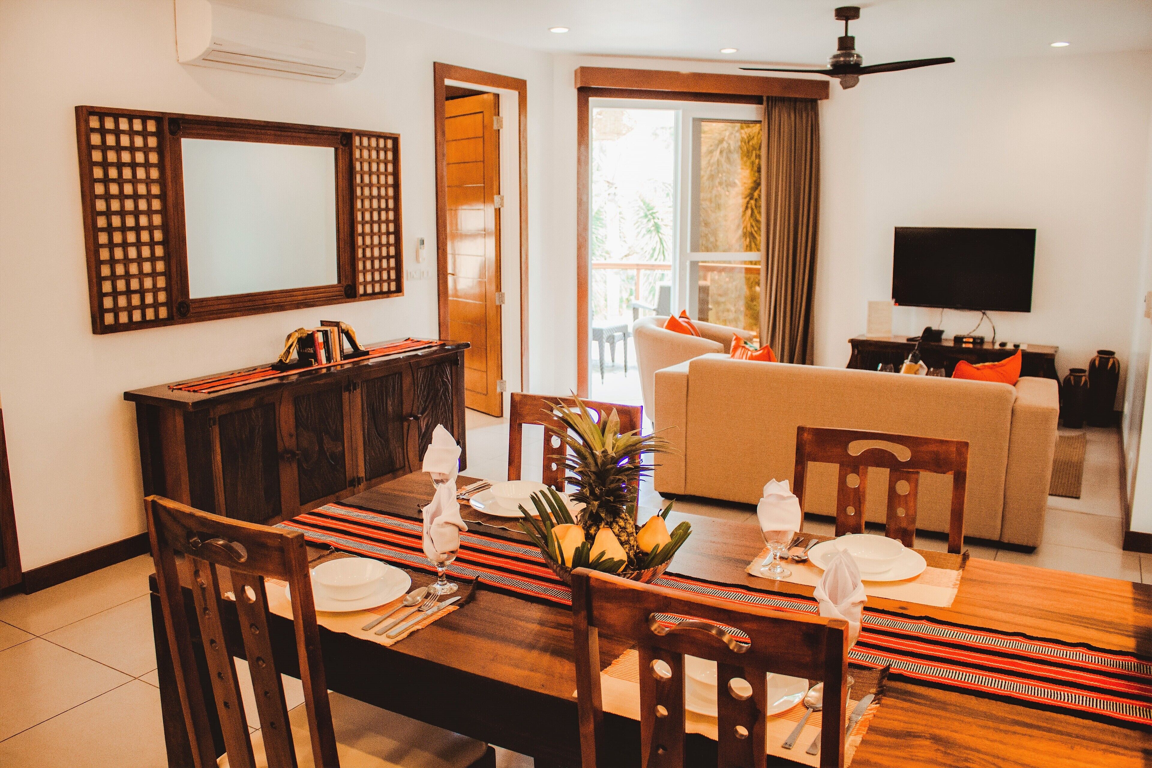 A dining area with a wooden table set for four, a sofa in the background, and a TV mounted on the wall.