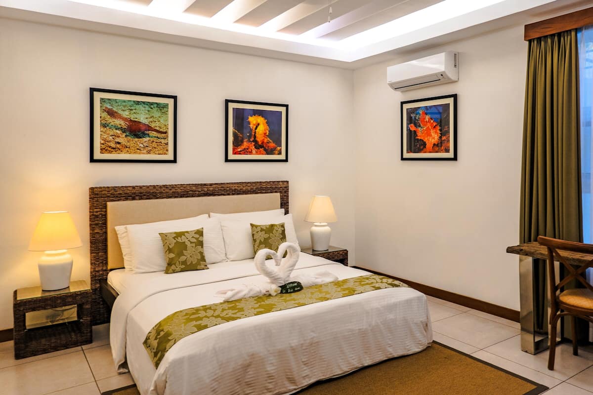 Une chambre d’hôtel comprenant un lit, deux tableaux accrochés au mur, une table de chevet avec une lampe, une chaise en bois et un appareil de climatisation.
