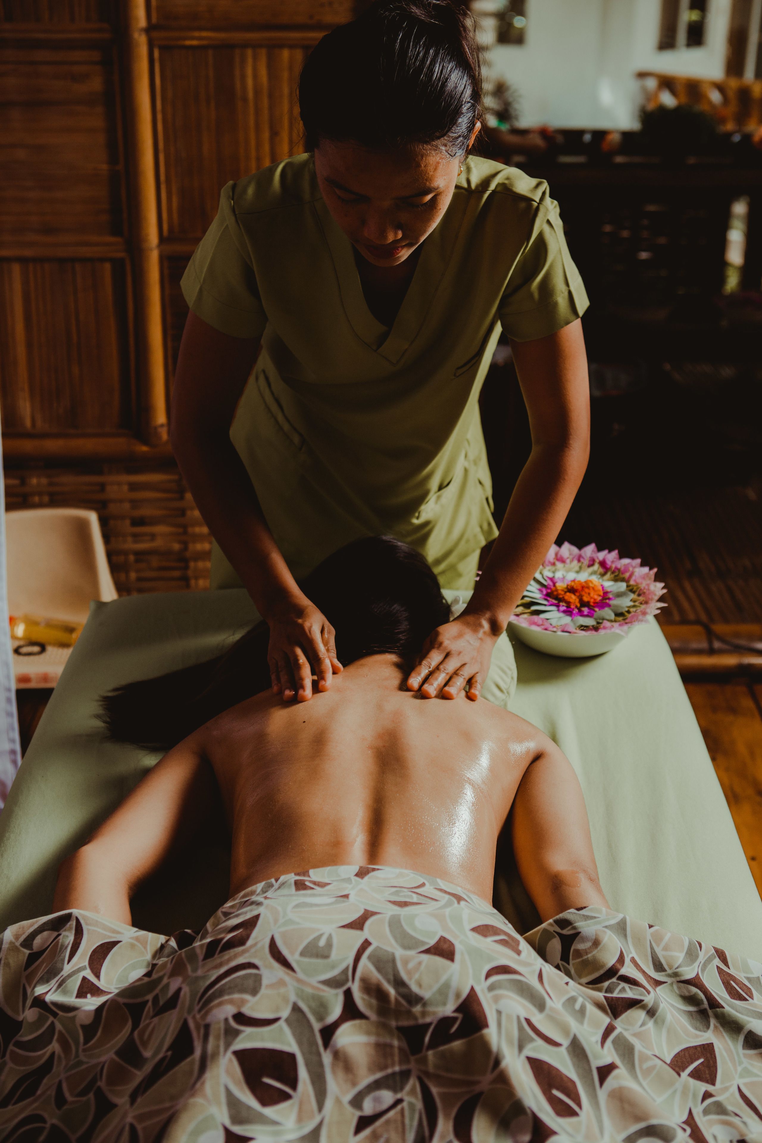 A person receiving a back massage in a room with wooden paneling and a bowl of flowers on a table.