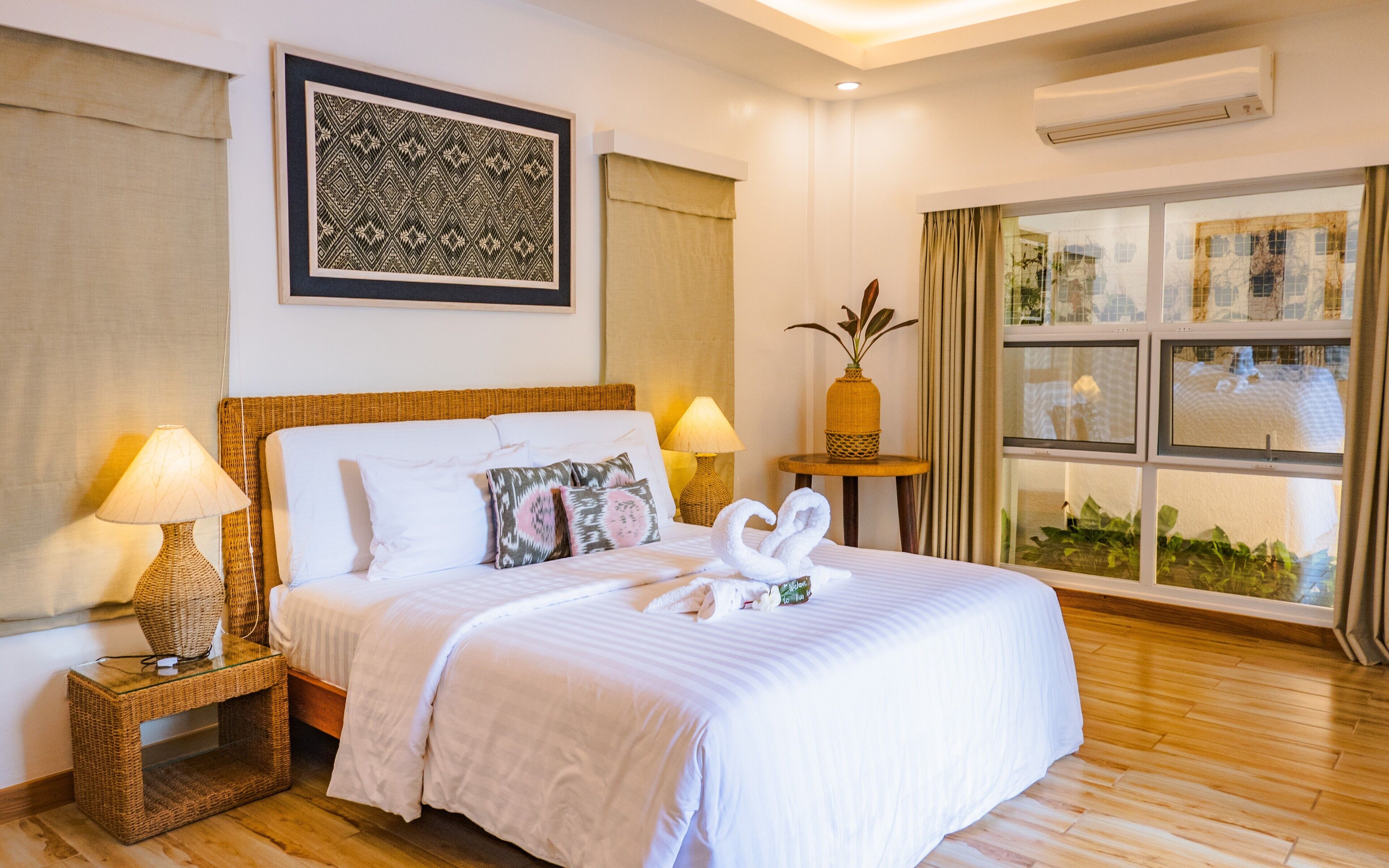 Una habitación de hotel con una cama grande, dos lámparas de noche, una obra de arte enmarcada sobre la cama, una ventana con cortinas y una mesita con un jarrón.