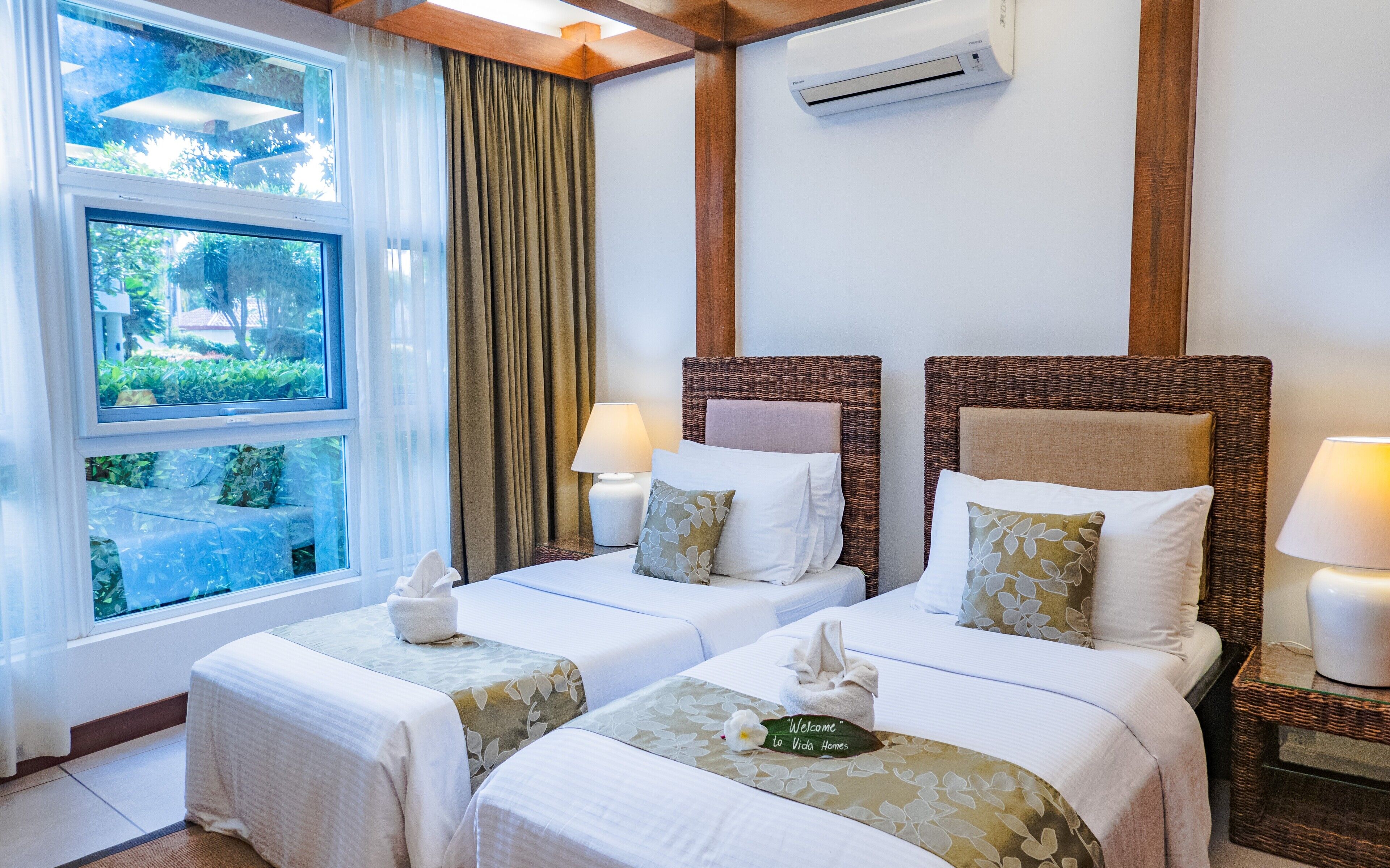 A hotel room with two beds, a window with curtains, a wall-mounted air conditioner, and a bedside table with a lamp.