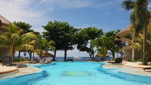 Une piscine d’un bleu éclatant, bordée de chaises longues et entourée de palmiers, le tout niché au milieu de bâtiments aux toits de chaume.