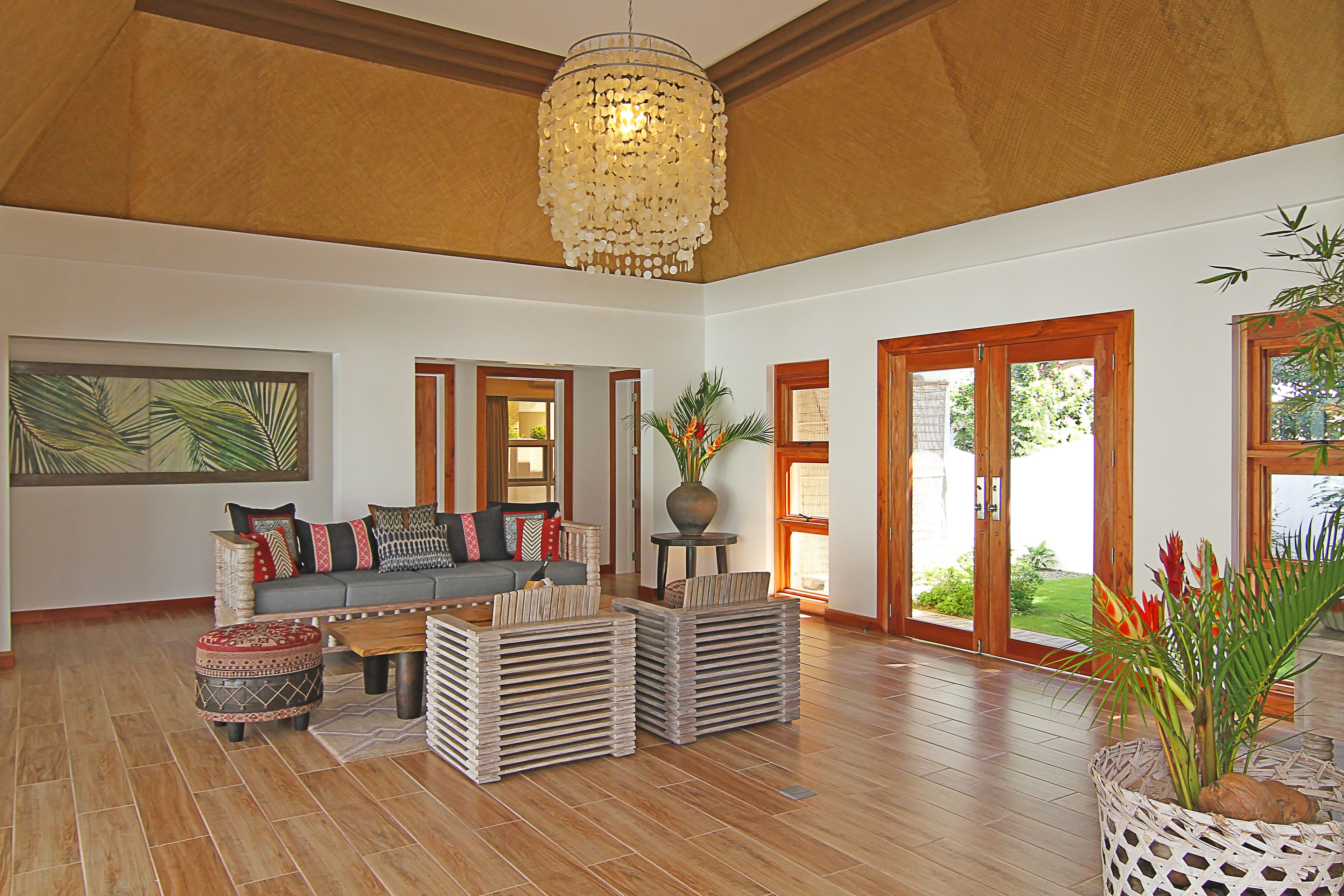 A spacious living room with a wooden ceiling, a large chandelier, a sofa set, and a view through the glass doors.