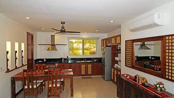 A modern kitchen with wooden cabinets, a dining table with chairs, a ceiling fan, and a large window with a view of greenery.