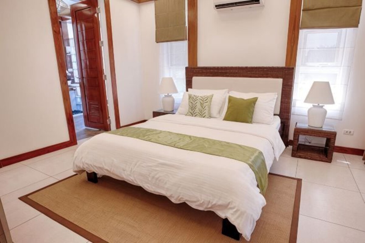 Un dormitorio con una cama grande, dos mesitas de noche con lámparas, un cabecero de madera y vistas al exterior a través de una ventana.