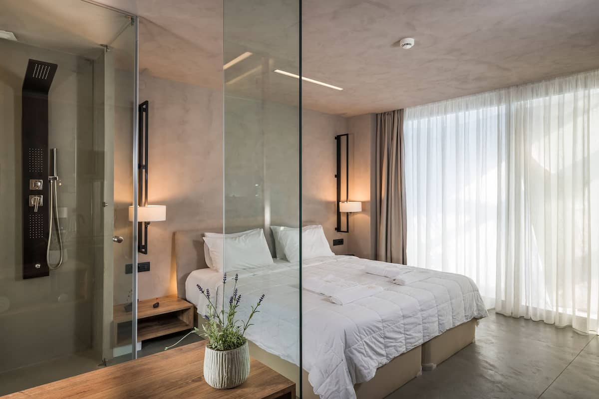 Una habitación de hotel moderna con una cama grande, cabina de ducha de vidrio y vistas al exterior a través de cortinas translúcidas.