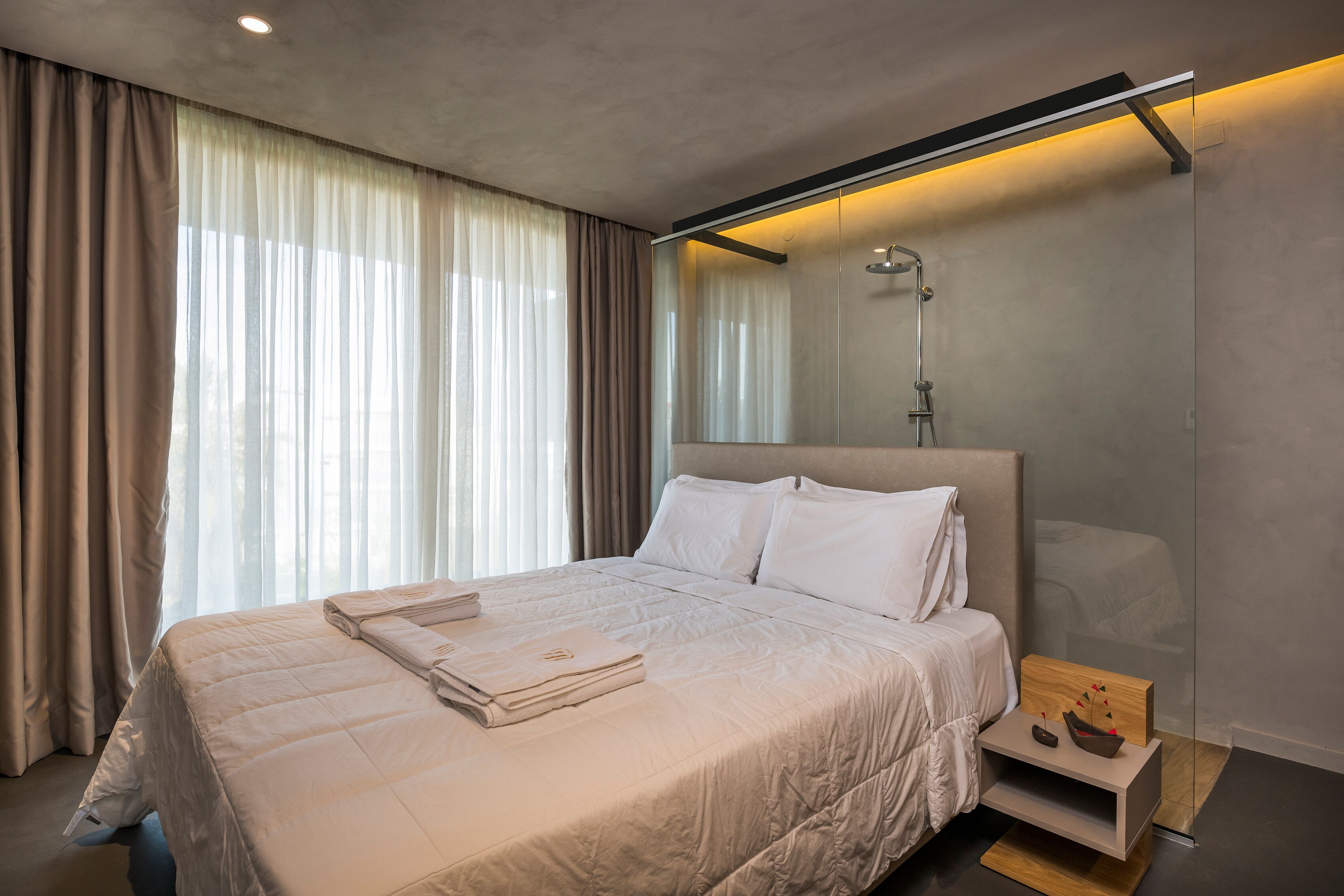 A modern hotel room with a large bed, a bedside table with a lamp, and a view through a window with sheer curtains.