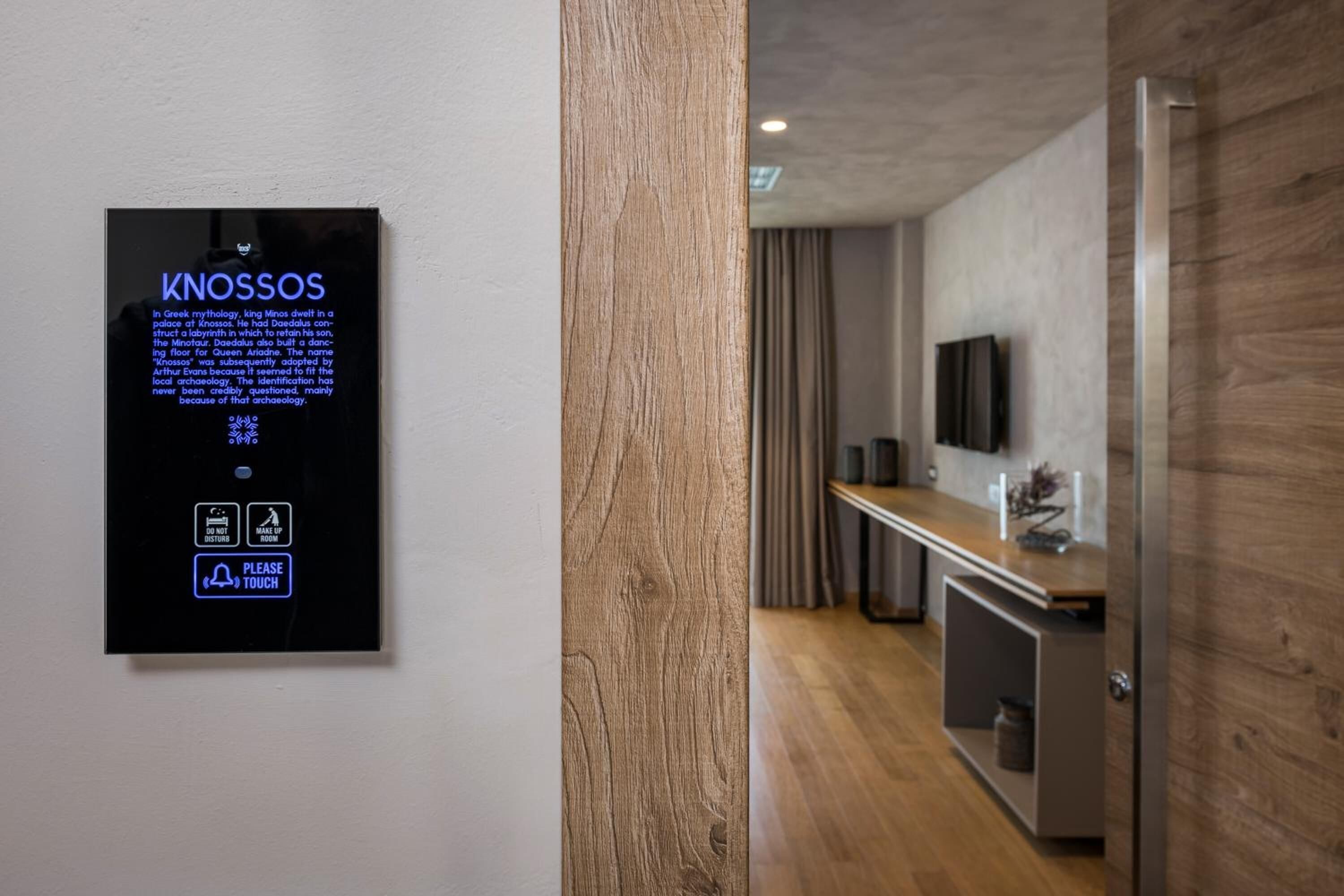 A modern hotel room with a wooden door, a mounted information board, a flat-screen TV, and a kitchenette area.