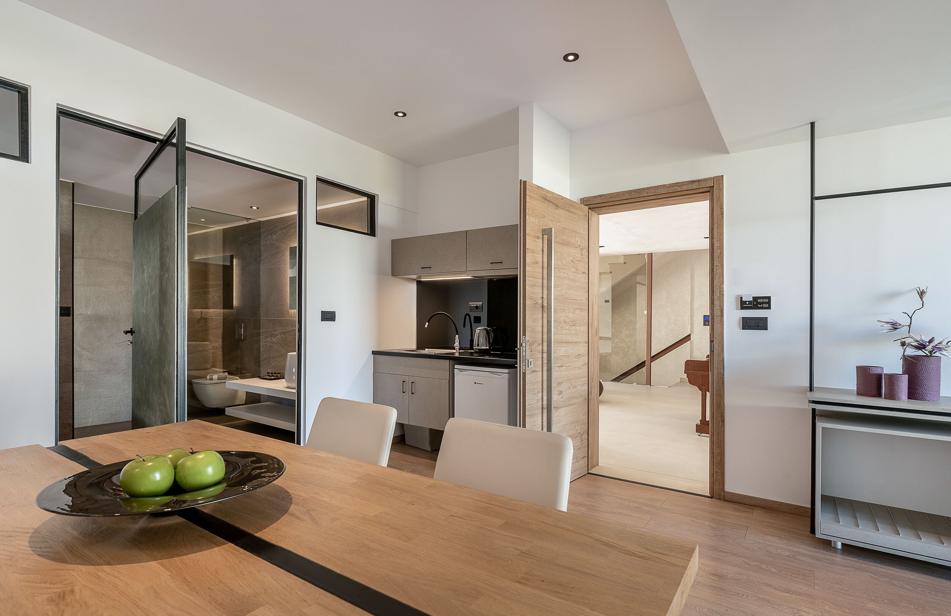 A modern kitchen with a dining area, featuring a wooden table with a bowl of apples, white chairs, and a view into the adjacent living space.