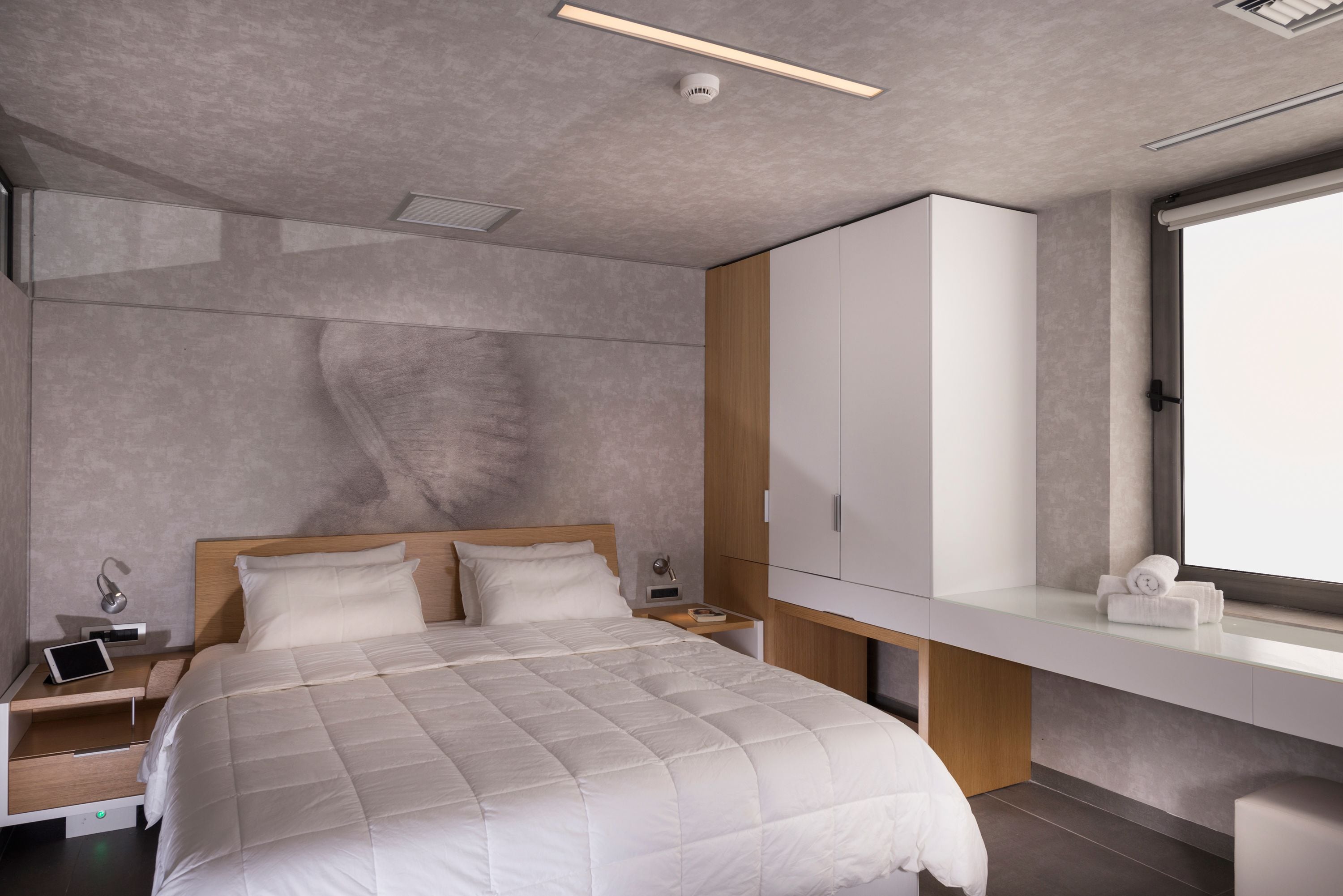 A modern bedroom with a large bed, a bedside table with a lamp, a wardrobe, and a window with a view.