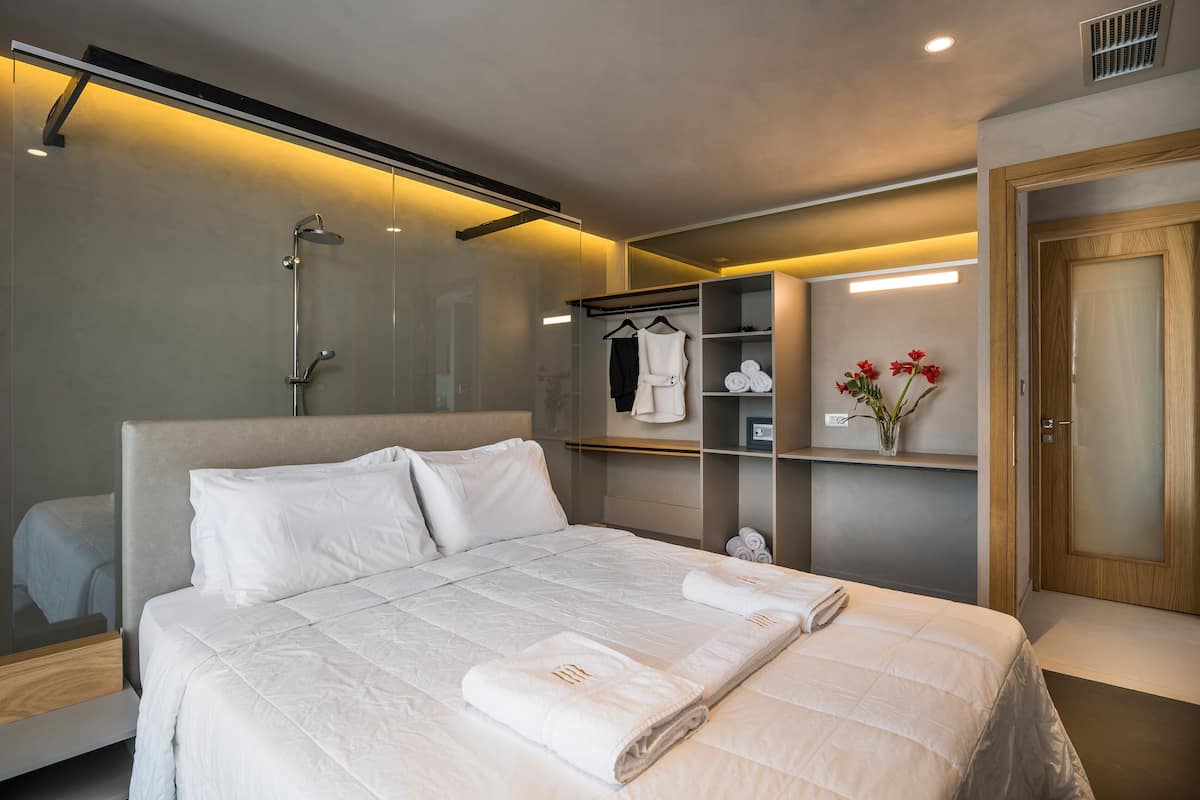 Una habitación de hotel moderna con una cama grande, ducha integrada, tocador con espejo y armario con batas.