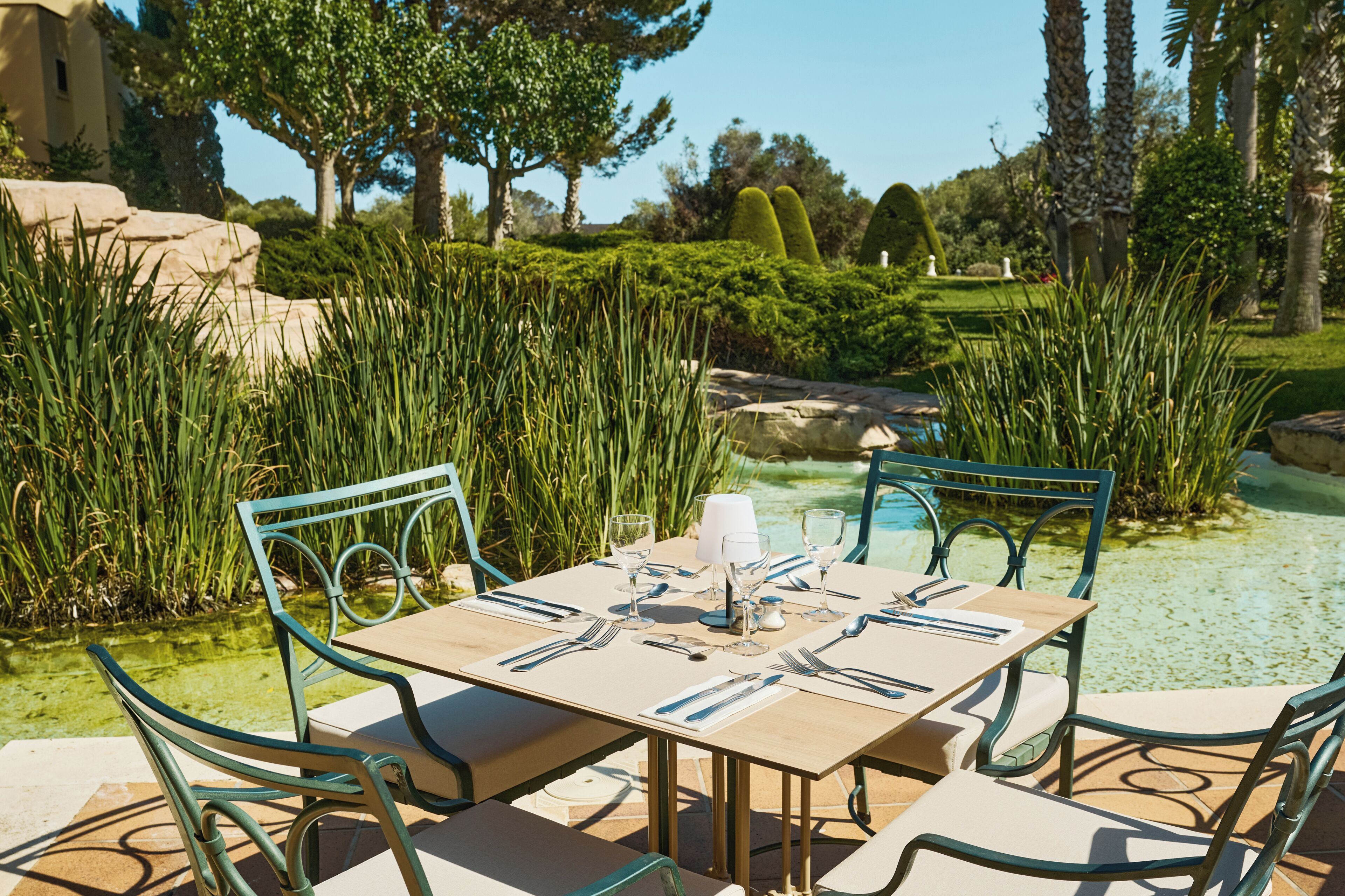 Un área de comedor al aire libre con una mesa lista para dos personas, rodeada de exuberante follaje y una vista despejada a una piscina.