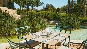 Un área de comedor al aire libre con una mesa lista para dos personas, rodeada de exuberante follaje y una vista despejada a una piscina.