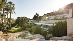 A landscaped garden with a pond, palm trees, and a building with outdoor seating. - Grupotel Playa Club (Ciutadella de Menorca)