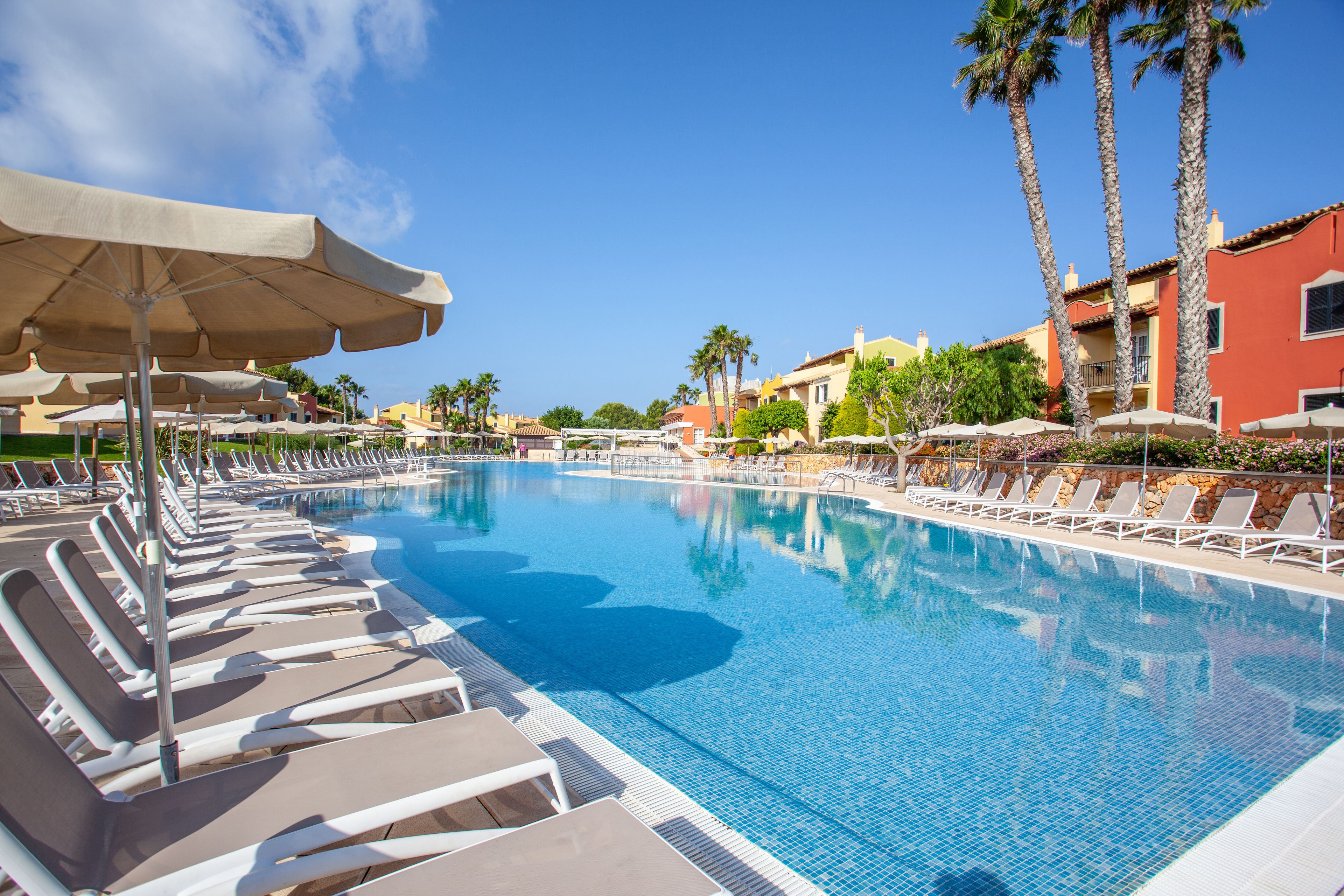 A swimming pool with lounge chairs and umbrellas, surrounded by palm trees and buildings.