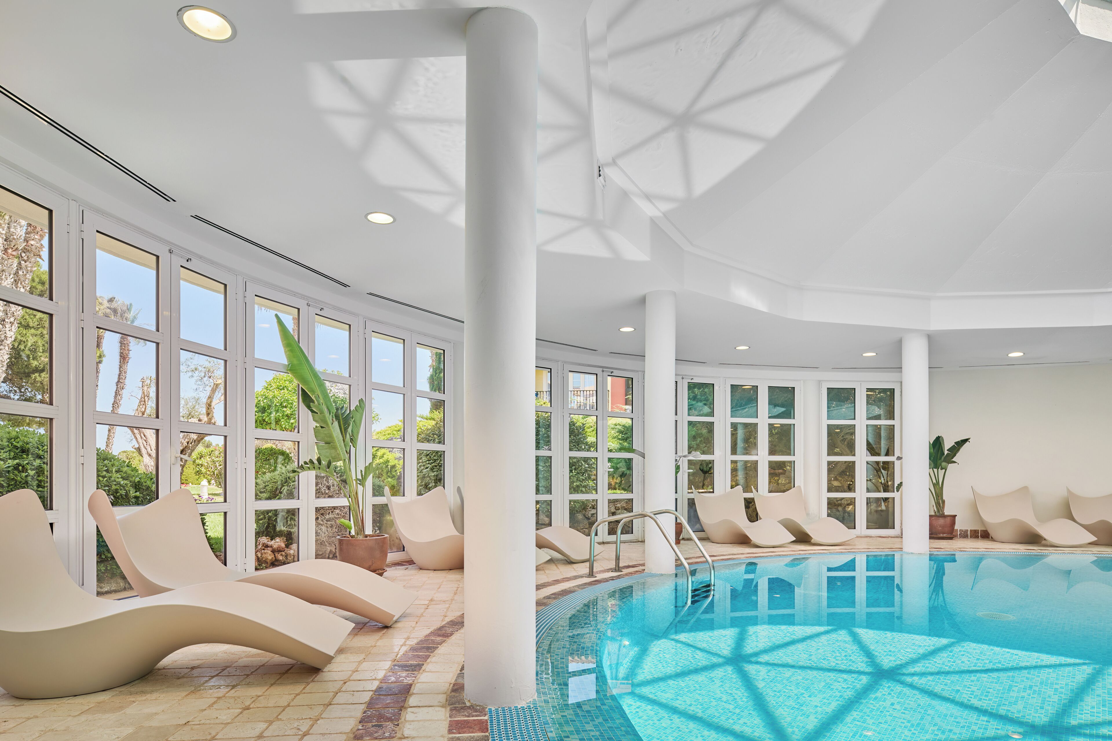 Una piscina interior moderna con paredes de vidrio transparente, múltiples sillones de descanso y suelo de baldosas.