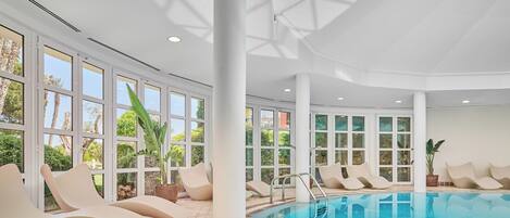 Una piscina interior moderna con paredes de vidrio transparente, múltiples sillones de descanso y suelo de baldosas.