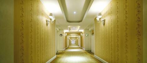 A hotel hallway with patterned wallpaper, wall sconces, and a carpeted floor.