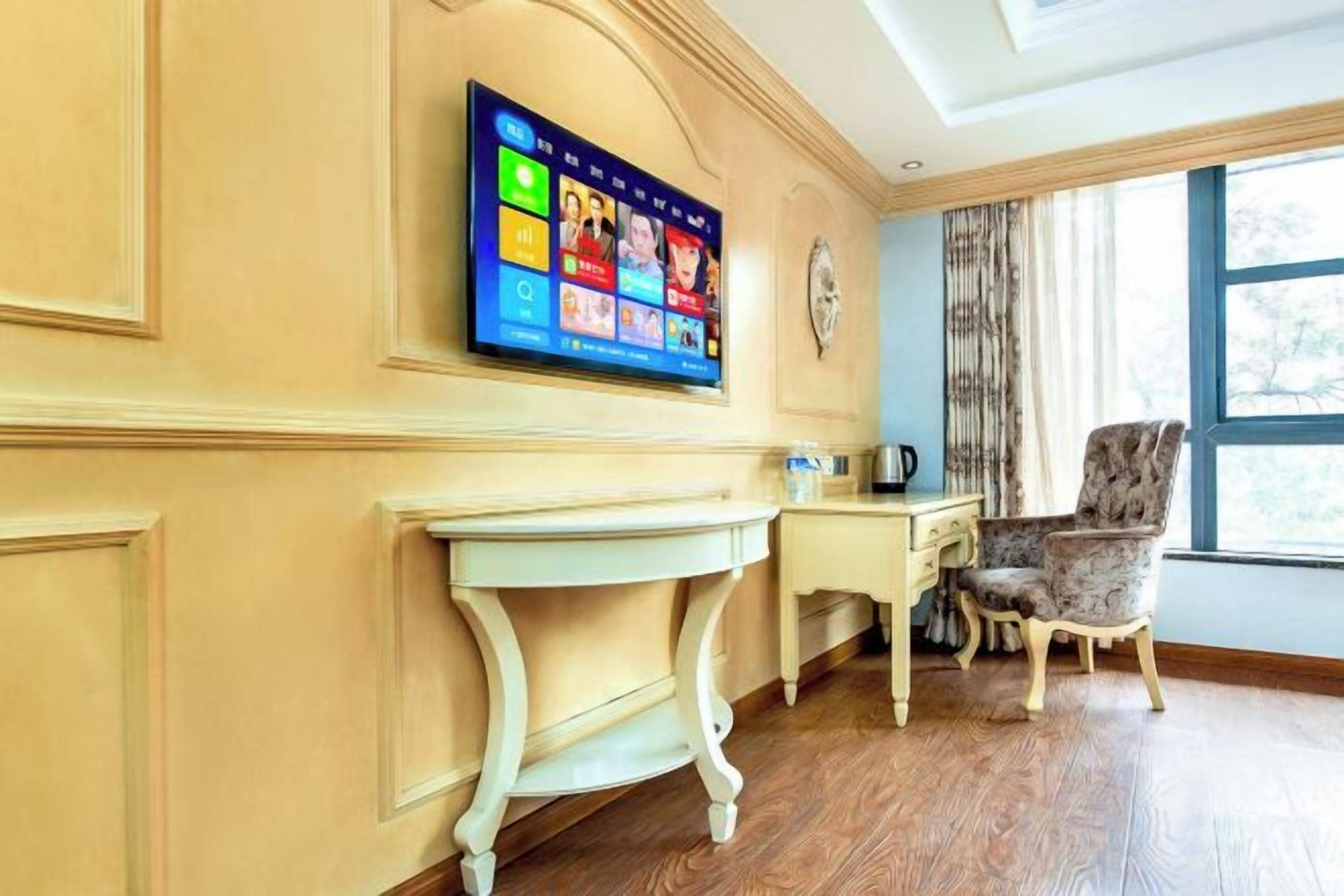A room with a flat-screen TV mounted on the wall, a small table, a chair, and a window with curtains.