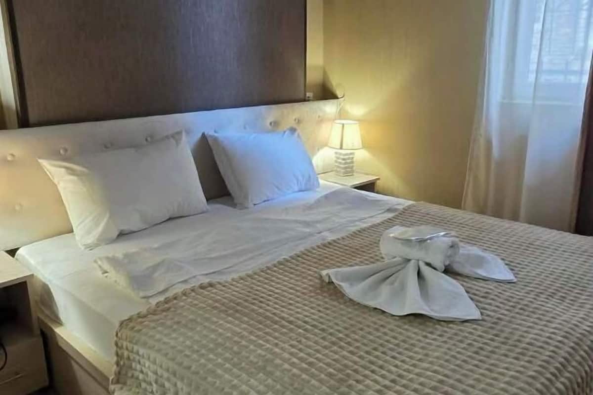 Una habitación de hotel con una cama, almohadas, una mesita de noche con una lámpara y una ventana con cortinas.