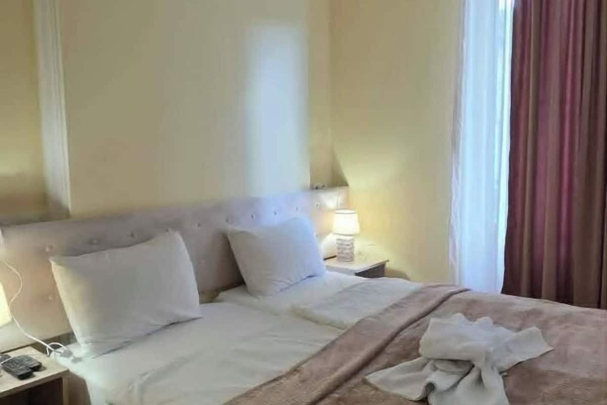 Habitación de hotel con cama, almohadas, mesita de noche con lámpara y ventana con cortinas.