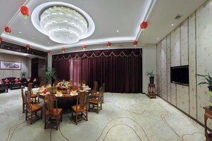 Un salón de banquetes con una mesa redonda dispuesta para comer, rodeada de sillas, una gran lámpara de araña y un telón rojo de fondo.