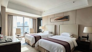 A hotel room with two beds, a desk with a chair, a window with a city view, and a painting on the wall. - Huacheng International Hotel (Lechang)