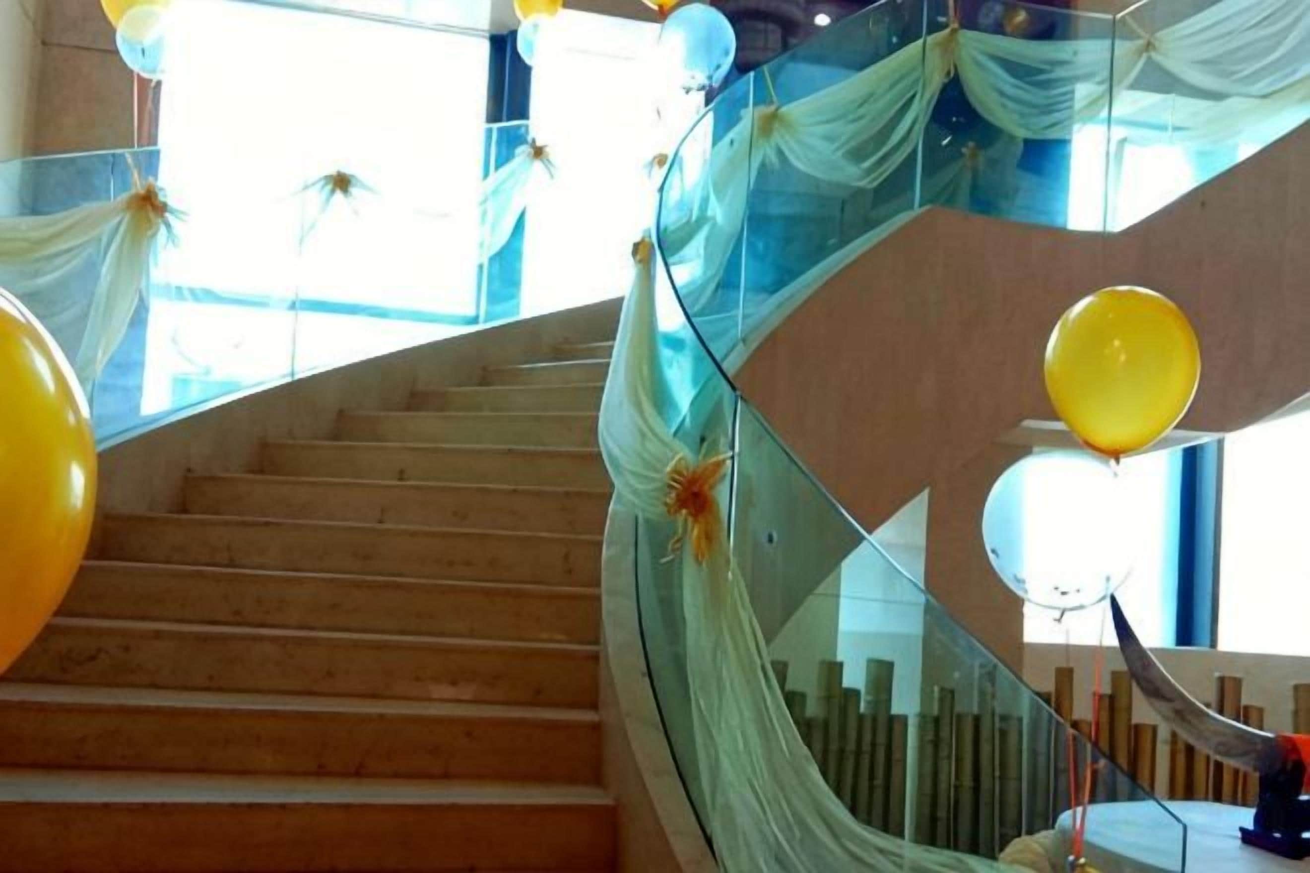 A spiral staircase with a glass railing, wooden steps, and yellow balloons.