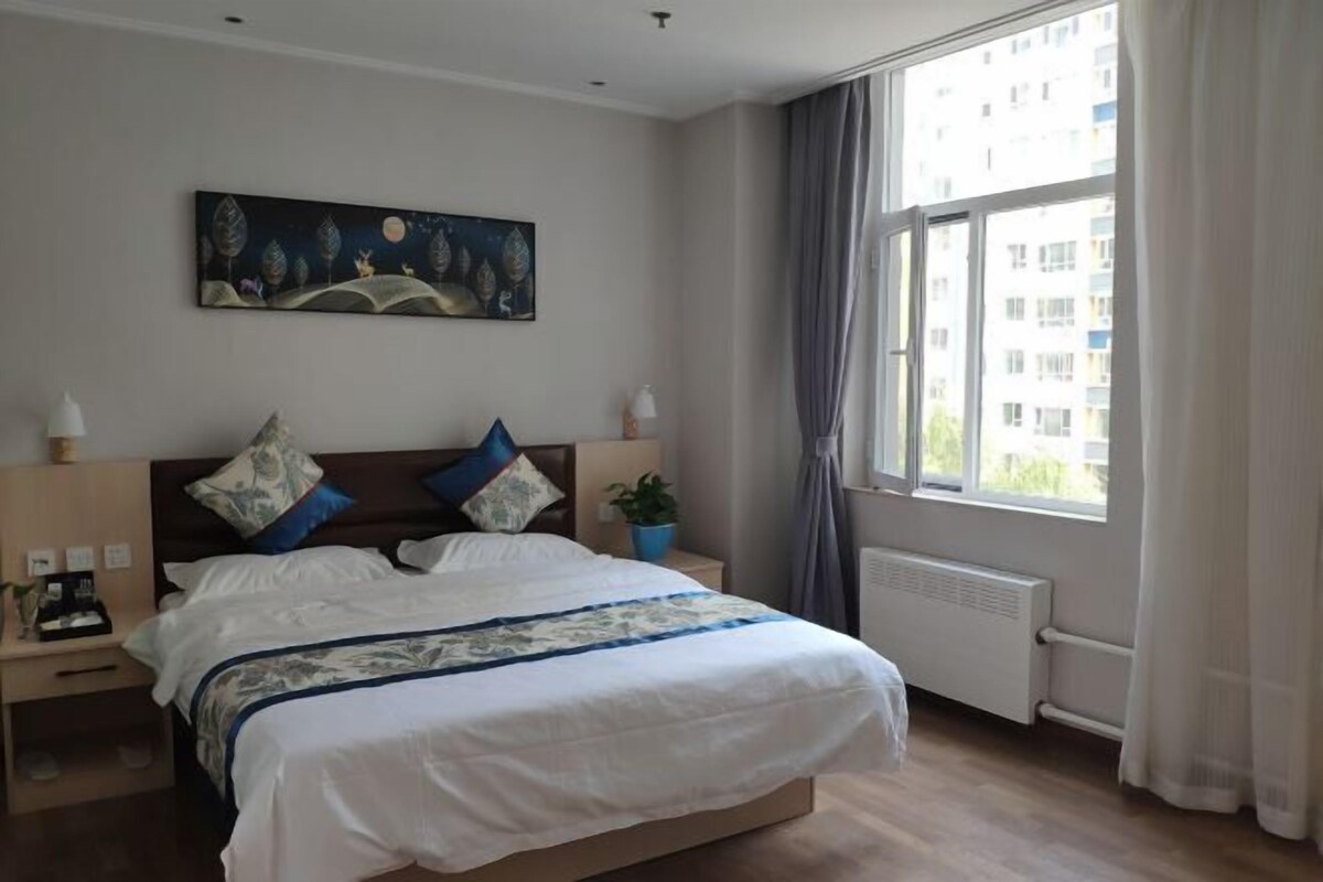 A bedroom with a bed, a nightstand, a lamp, a painting, a window with curtains, and a radiator.