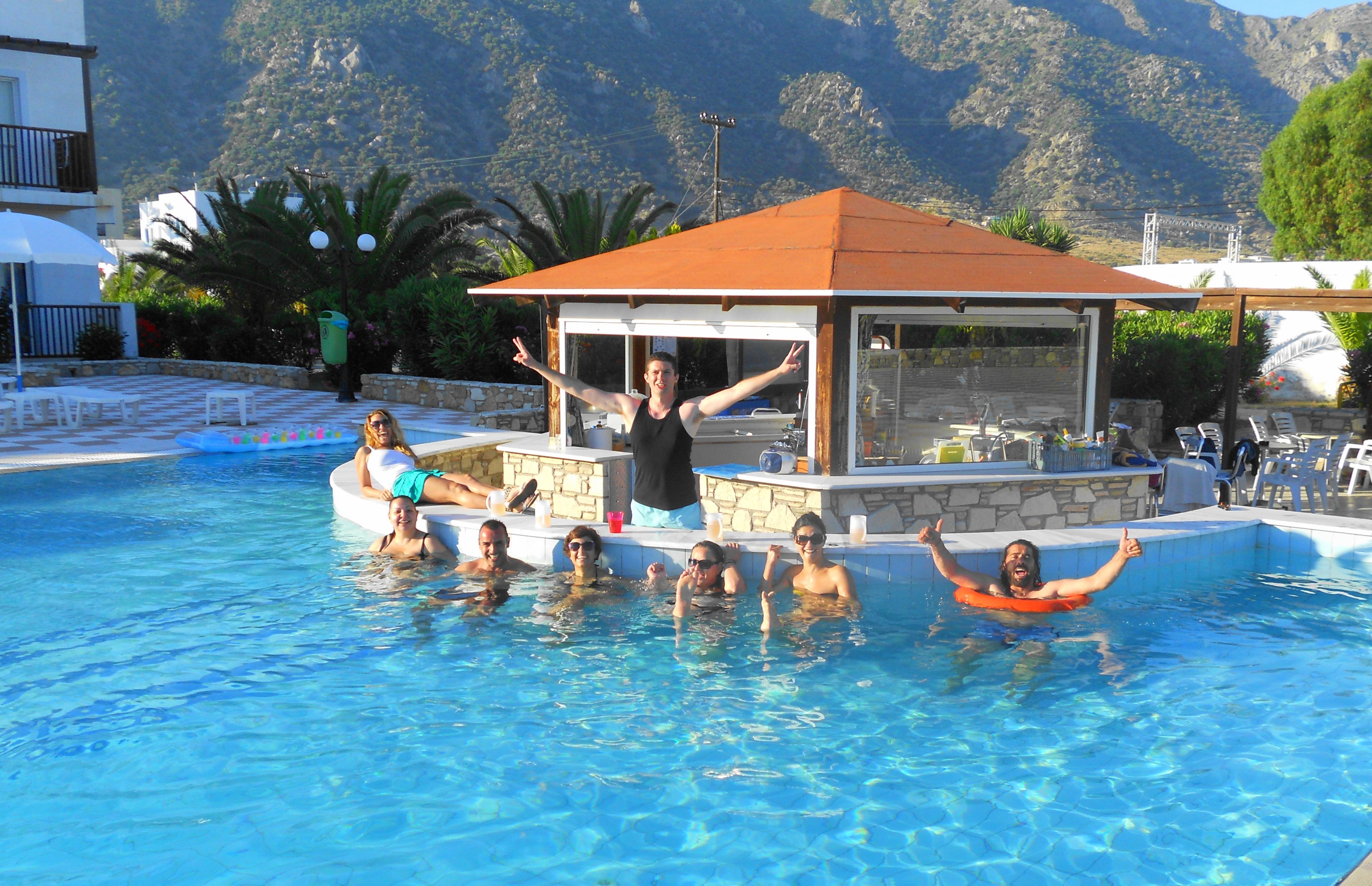 Ein Pool mit badenden Personen, ein überdachter Bereich mit Tischen und Berge im Hintergrund.
