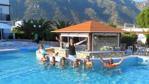 Una piscina con gente disfrutando en el agua, un área techada con mesas y montañas al fondo.