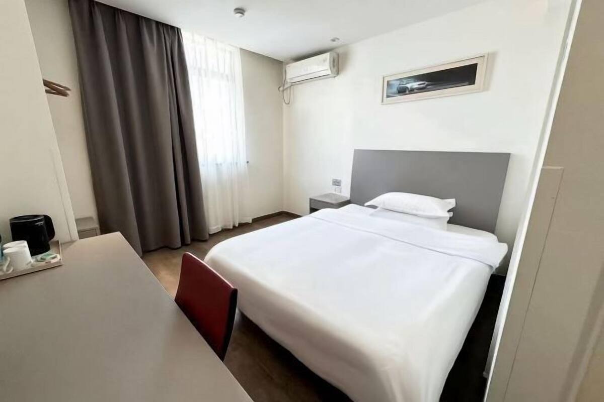 A hotel room with a bed, a desk, a chair, a nightstand, a lamp, a window with curtains, and an air conditioning unit.