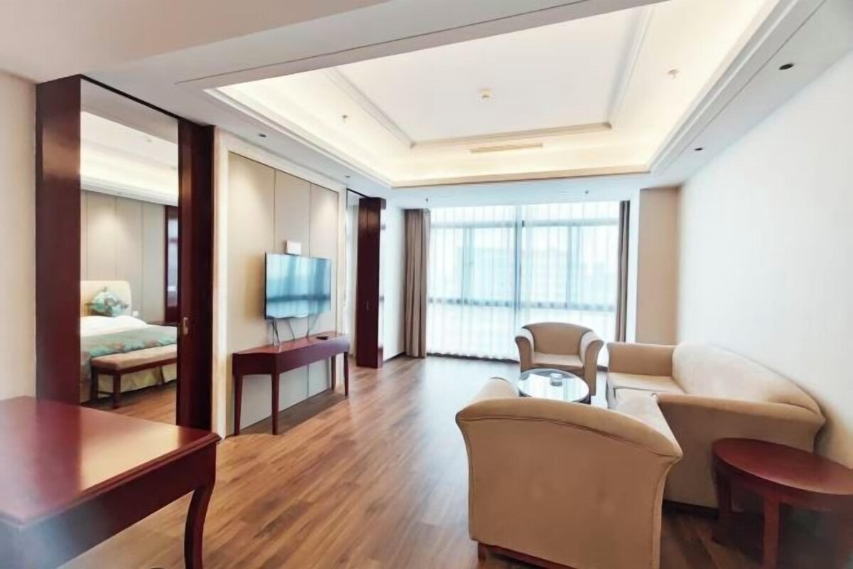 Quarto de hotel moderno com uma cama grande, televisão de tela plana, sofá, mesa de centro e vista para o exterior por grandes janelas.