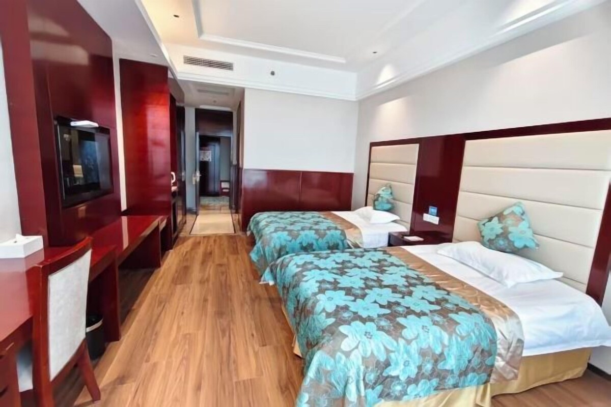 Quarto de hotel com duas camas, uma escrivaninha, televisão e banheiro visível por uma porta aberta.