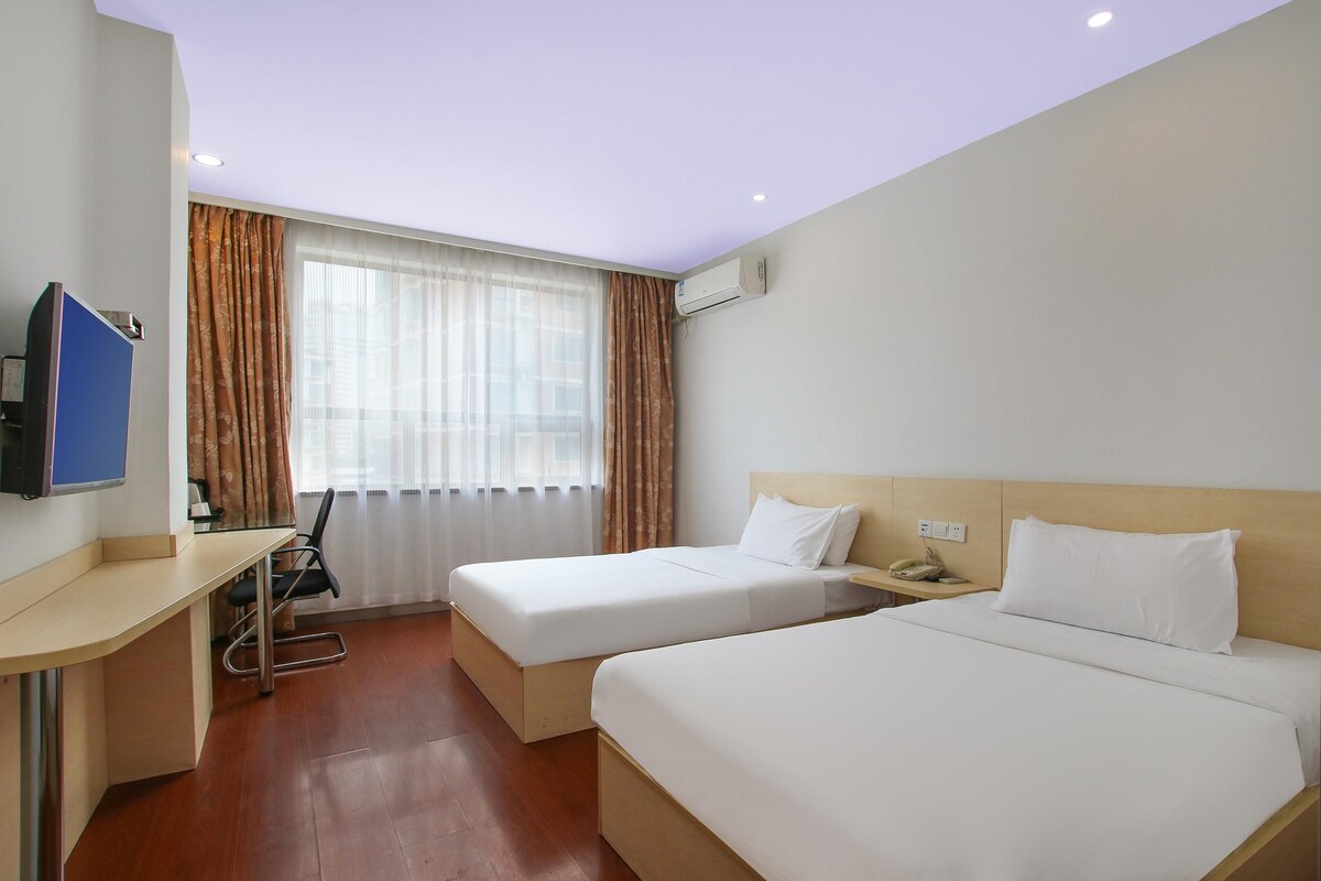 A hotel room with two single beds, a desk with a chair, a television, and an air conditioning unit.